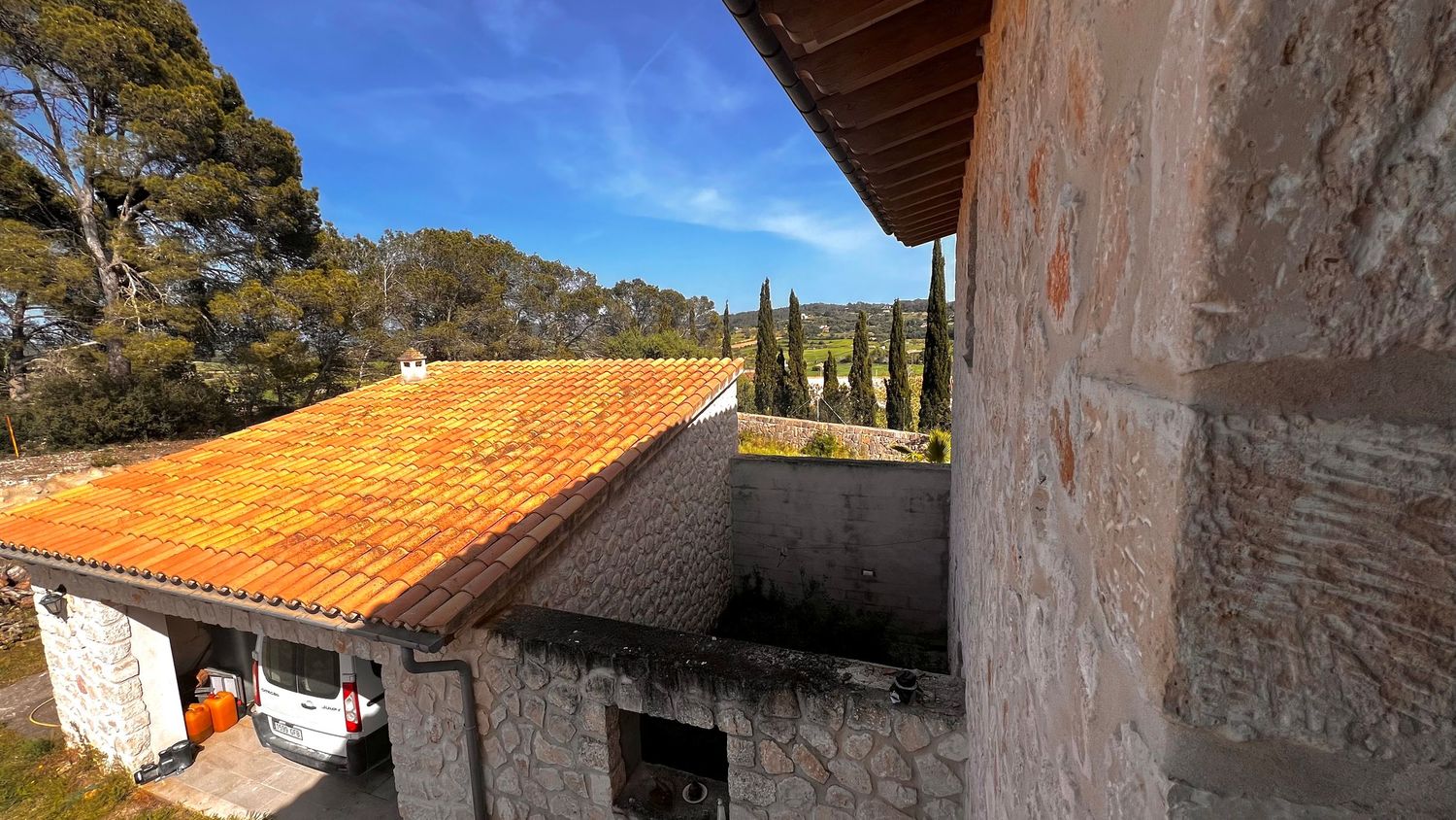 Stone building with orange terracotta roof tiles surrounded by cypress trees under a bright blue sky in a Mediterranean landscape.