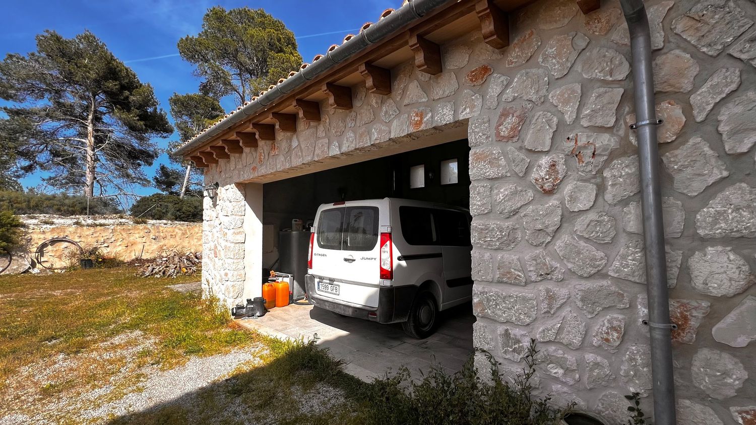Stone garage with open door housing a white van, surrounded by trees and dry grass in a rural setting.