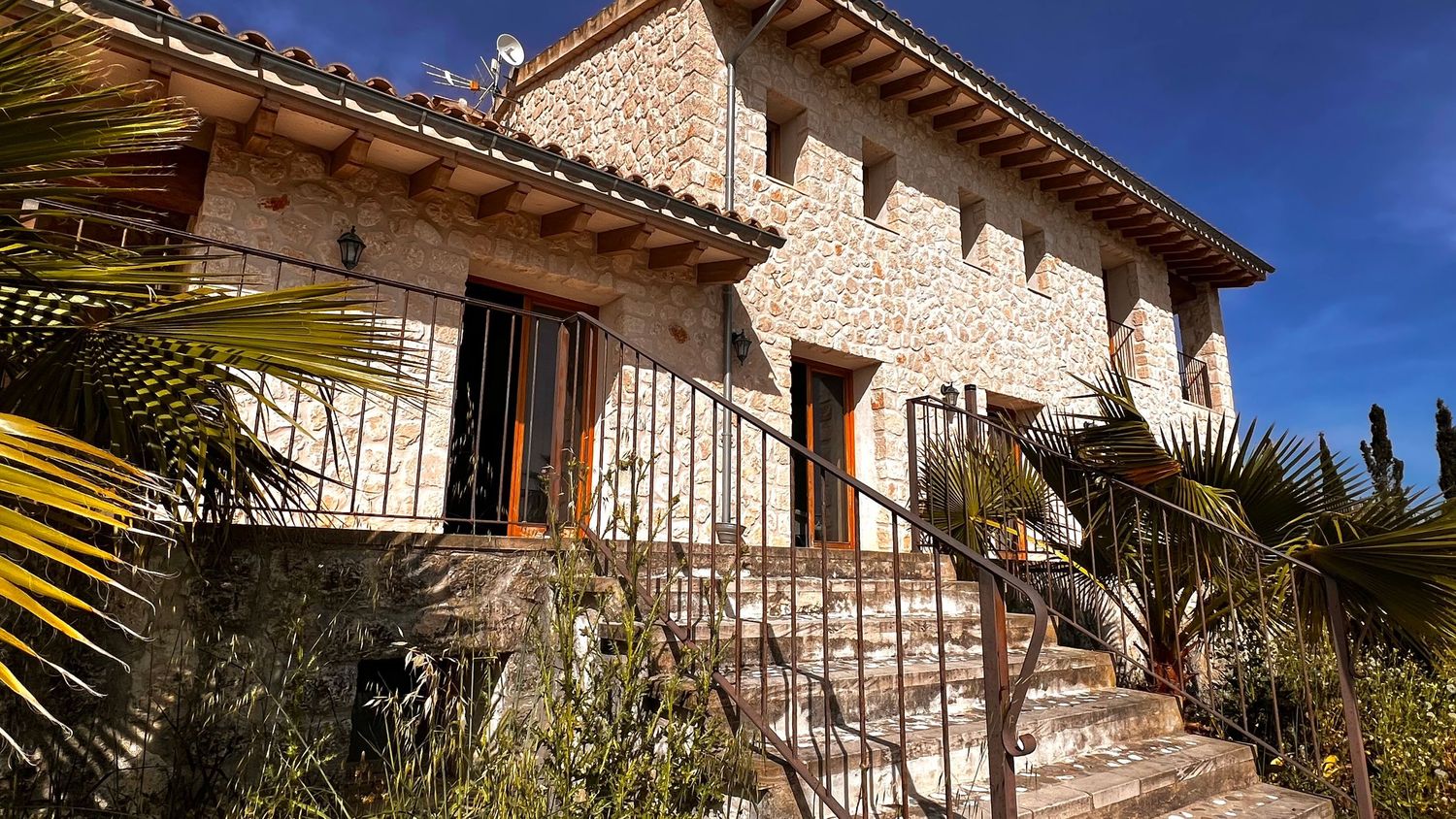 Stone villa with terracotta roof tiles, wooden shutters, palm trees, and stone staircase leading to entrance in sunny Mediterranean setting.
