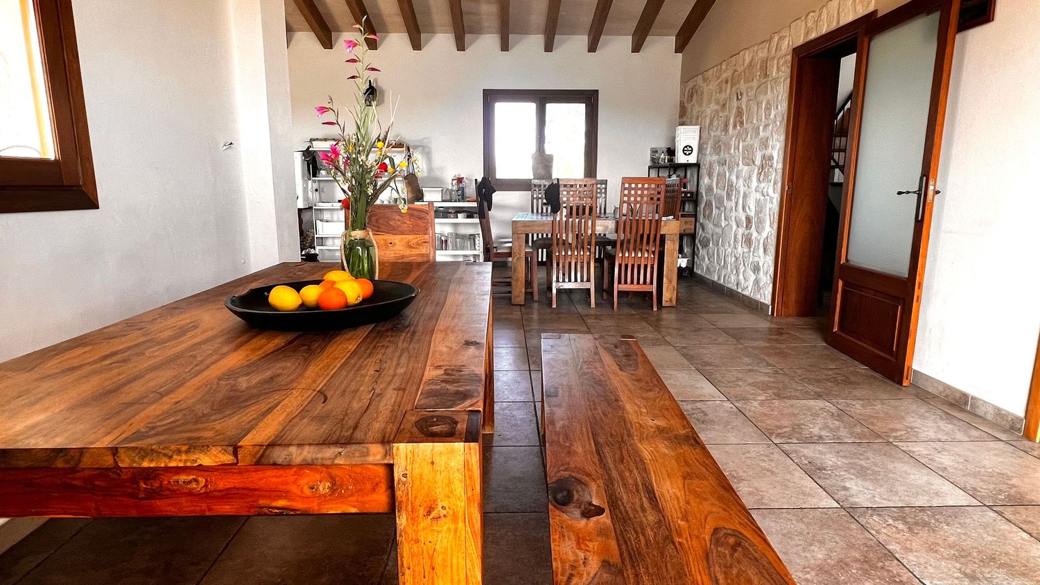 Rustic dining room with a large wooden table, stone wall, and tiled floors in a bright, open space.