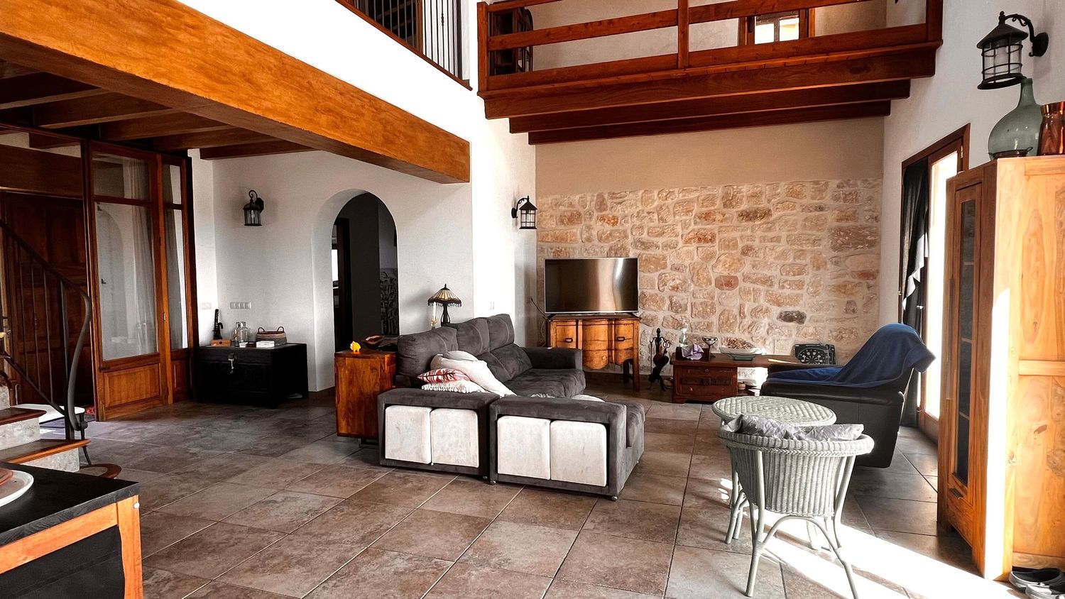Rustic living room with stone wall, tile floors, wooden beams, gray sofa, and dining area in a cozy Mediterranean-style home.