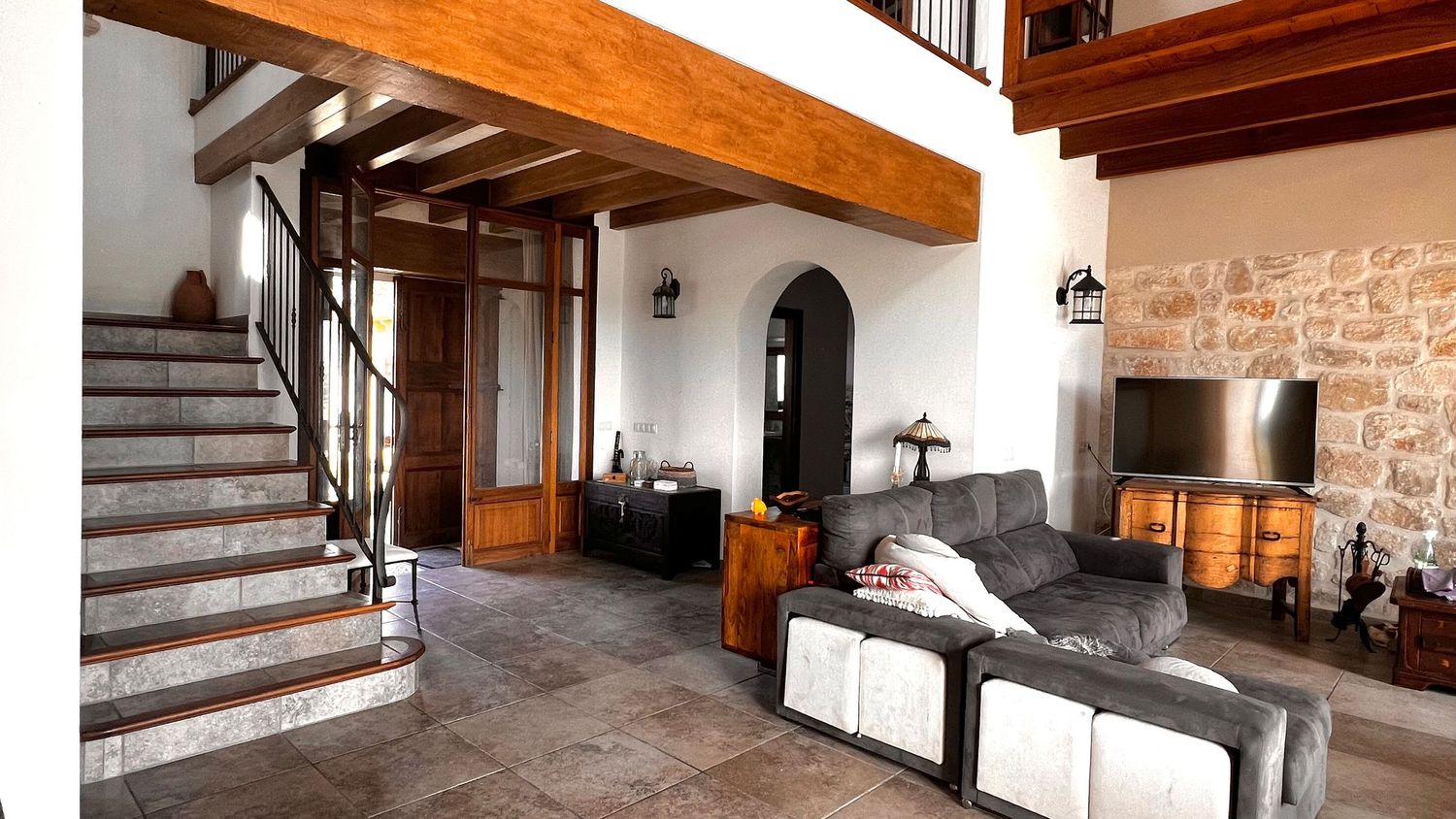 Rustic living room with stone walls, wooden beams, gray sectional sofa, TV unit, and a wooden staircase with wrought iron railing.