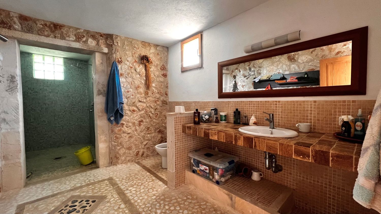 Rustic bathroom with stone walls, walk-in shower, mosaic tile floors, double vanity sinks, and a large mirror with warm lighting.