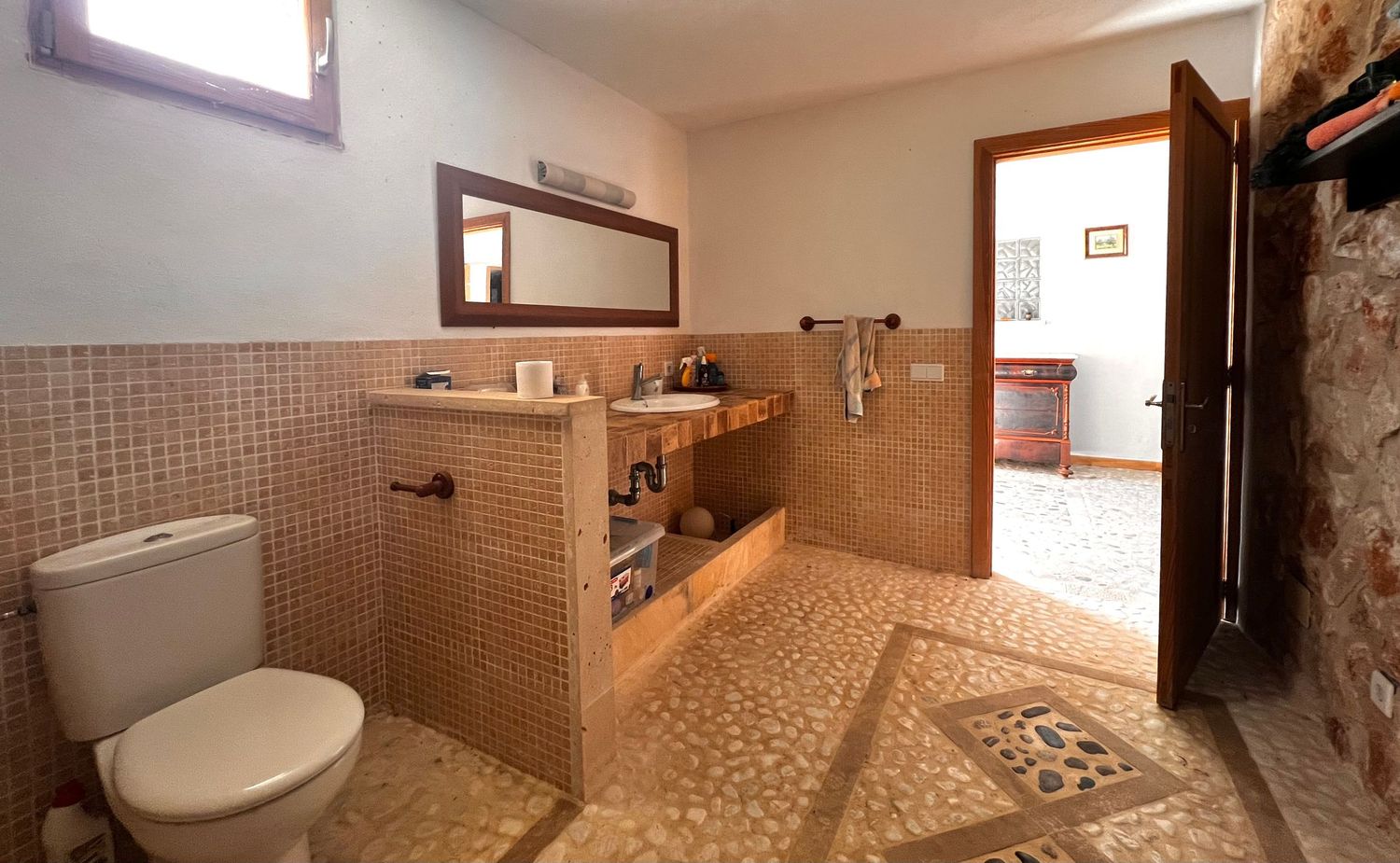 Rustic bathroom with mosaic tile floors, stone walls, wooden vanity, large mirror, and white toilet near an open wooden door.
