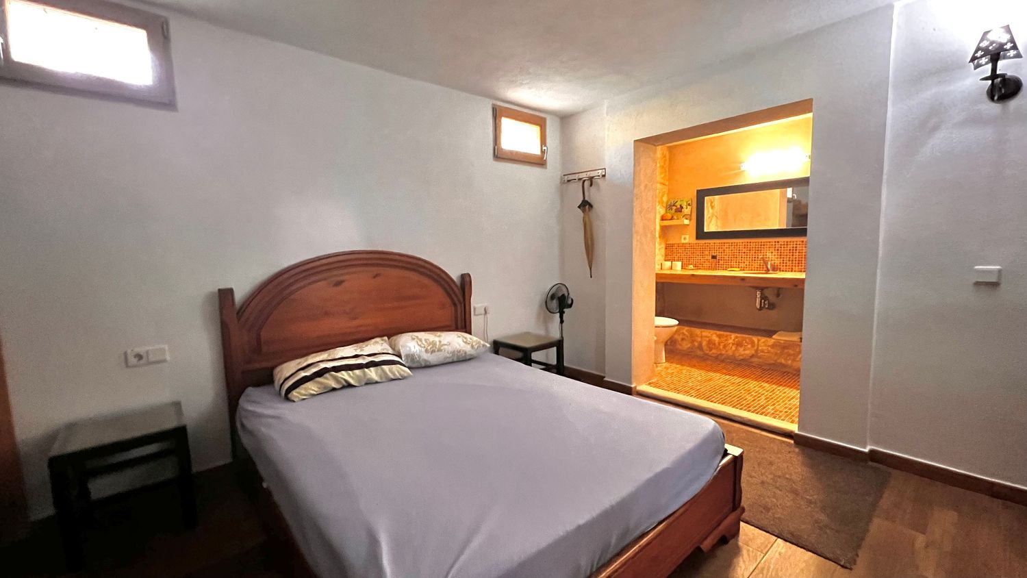 Cozy bedroom with wooden bed frame, dark hardwood floors, and an ensuite bathroom with warm lighting.