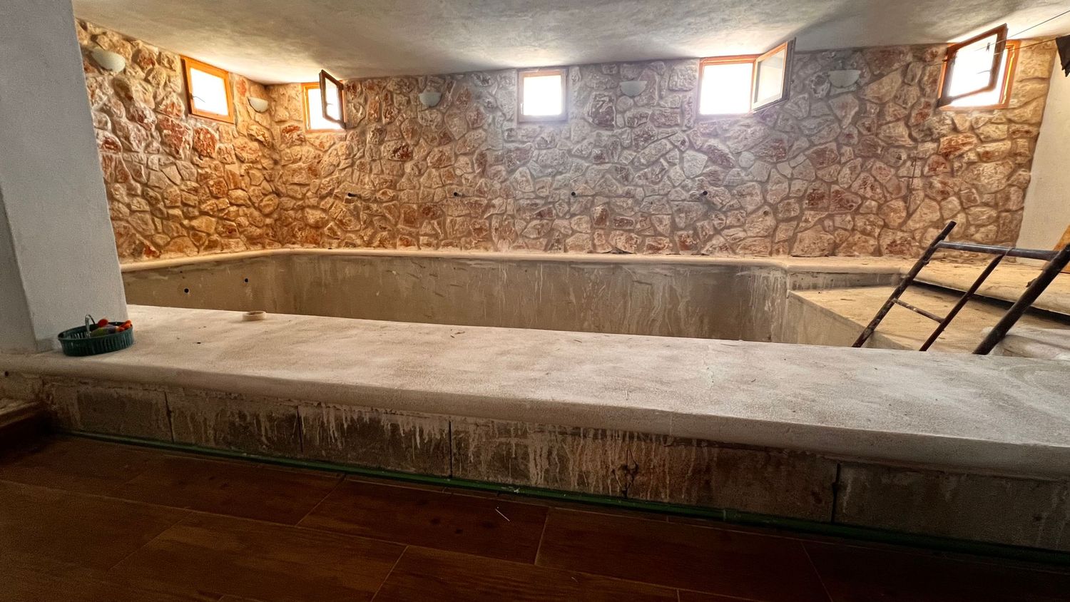 Ancient stone-walled underground pool with concrete basin, wooden flooring, small windows, and metal ladder in historic building.