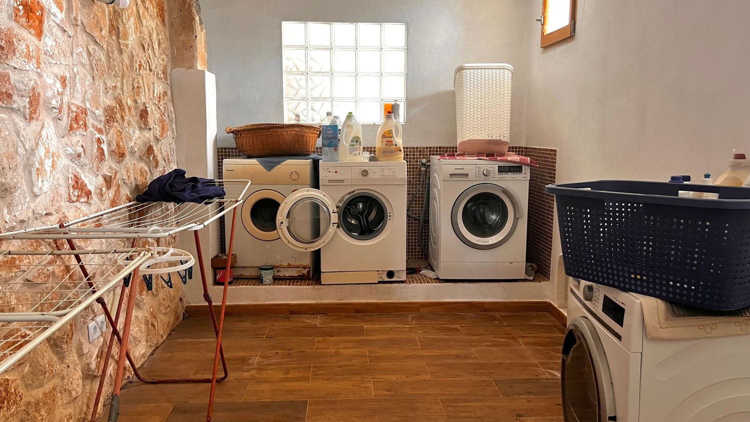 Laundry room with three washing machines, wooden floors, brick wall, drying rack, and laundry basket in a rustic space.