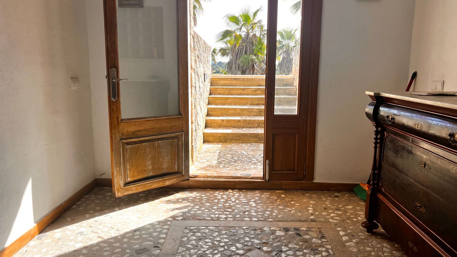Sunlit entryway with ornate tiled floor, wooden double doors opening to outdoor steps and palm trees beyond.