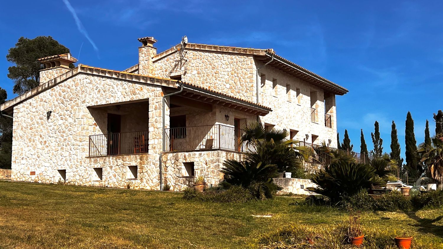 Large stone villa with balconies and terraces on a sunny hillside with cypress trees and blue sky.