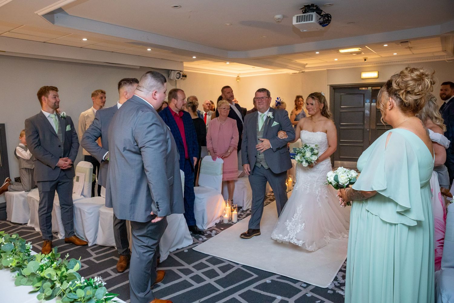 Bride walks down the aisle at an indoor wedding ceremony with guests standing and watching.