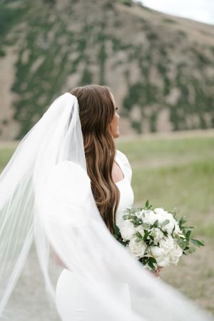 Bride with flowing veil holds white bouquet against mountain backdrop.