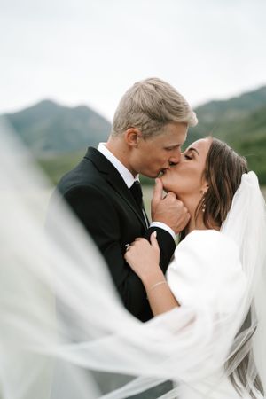 Intimate wedding kiss captured with flowing veil and mountain backdrop.