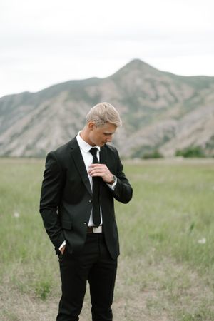 Groom in classic black suit adjusts tie while standing in mountain field.