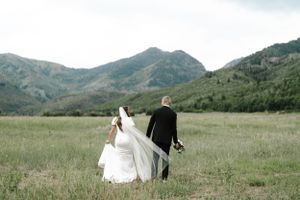 A wedding couple walks through a scenic mountain meadow with flowing white dress and veil.