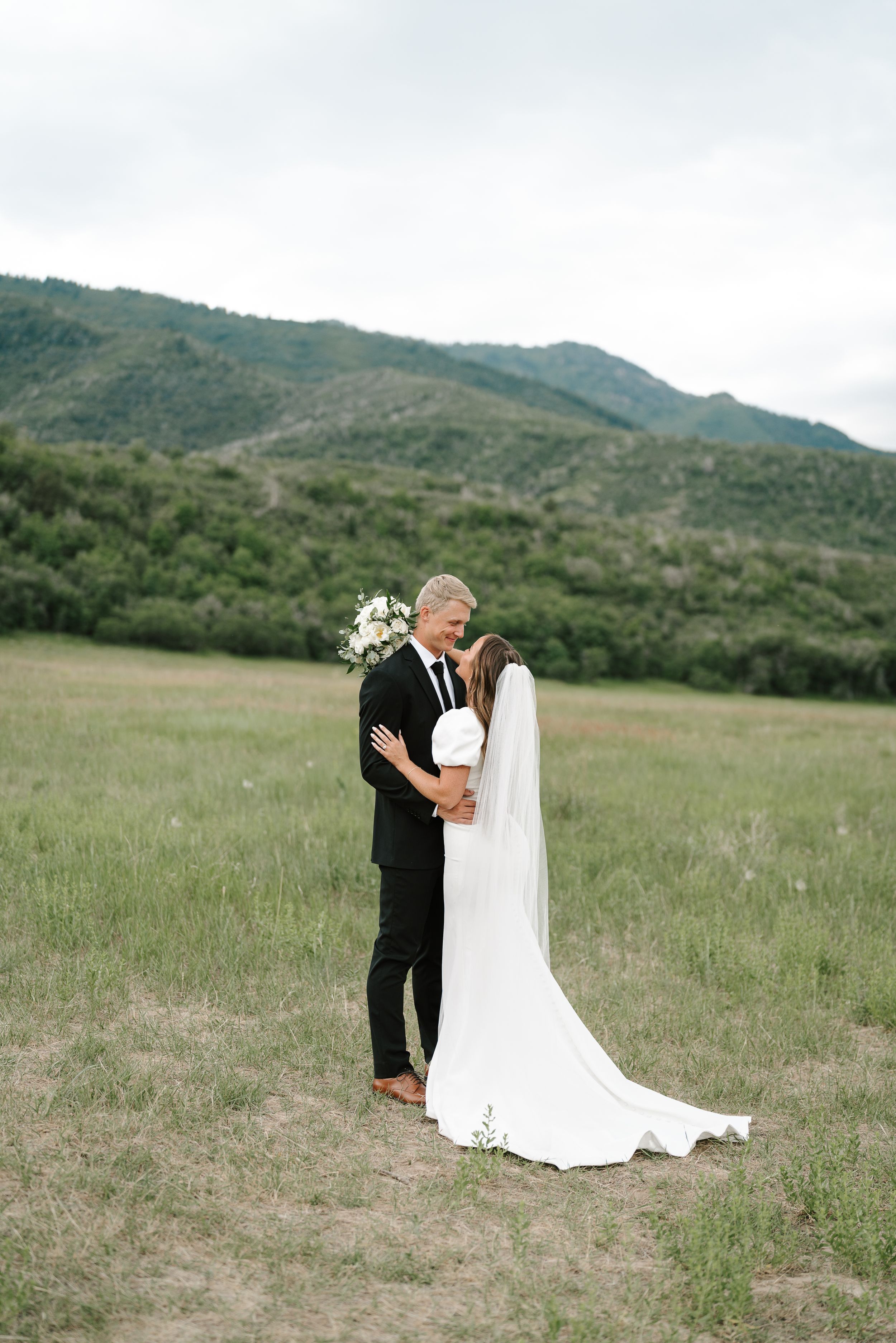 A romantic wedding portrait of a couple embracing in a mountain meadow with a flowing white gown and bouquet.