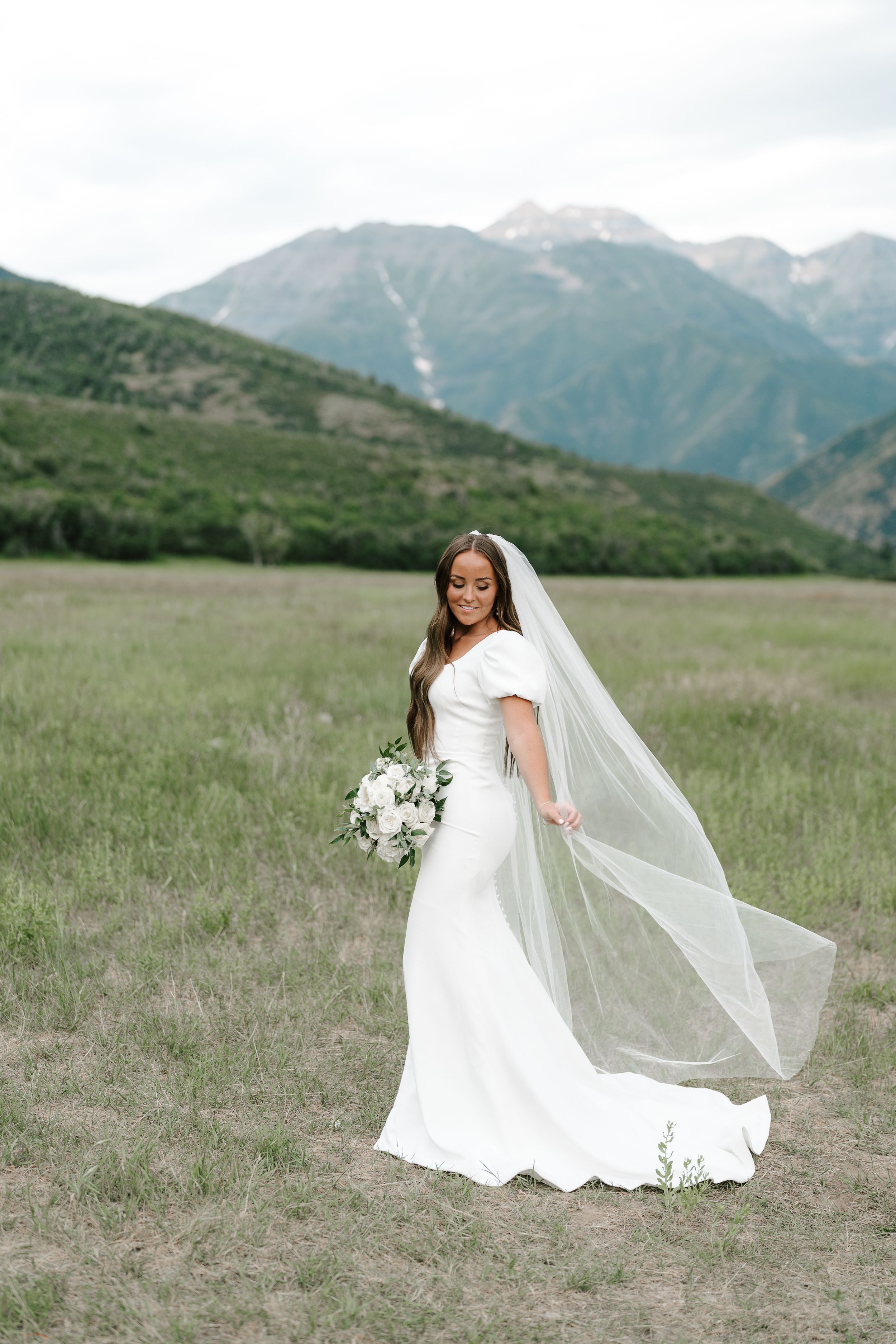 Bride in modern white dress with flowing veil poses in mountain meadow holding white bouquet.