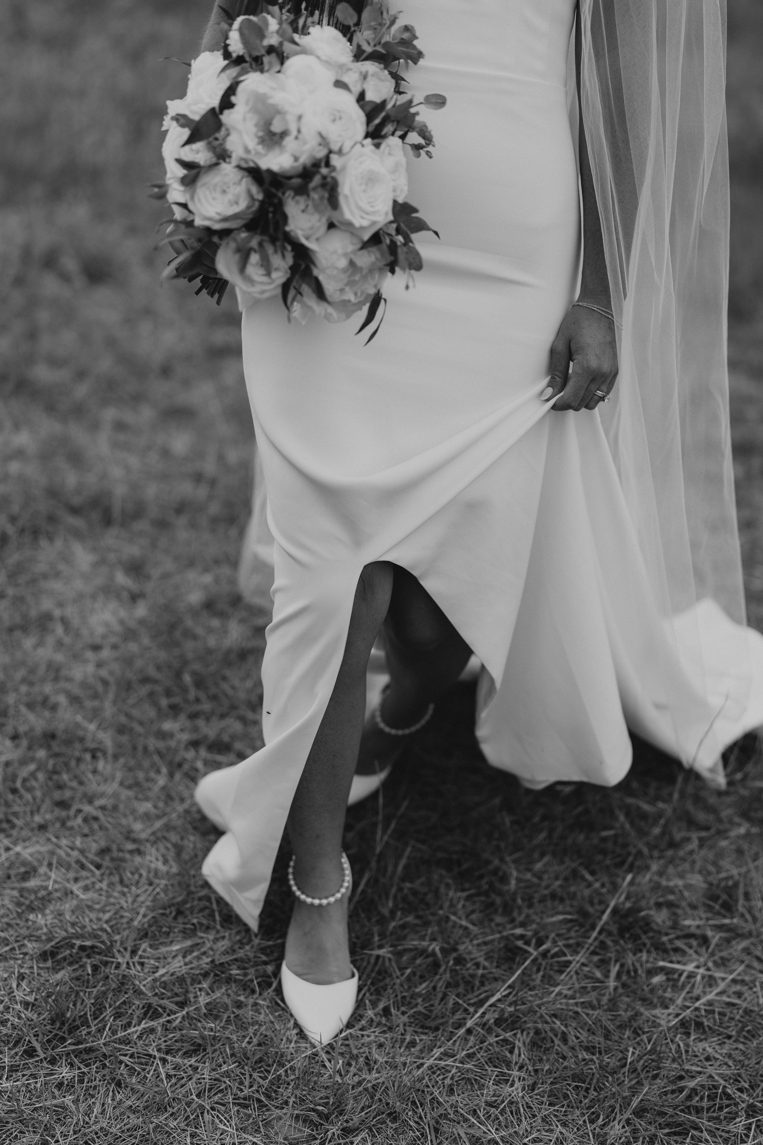 Black and white detail shot of bride's elegant white shoes and dress hem with bouquet.
