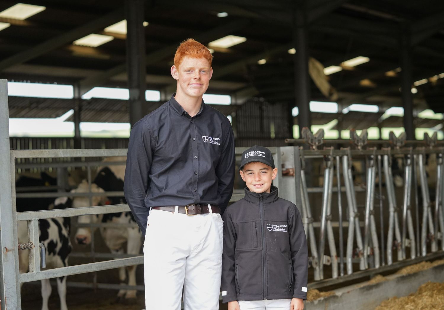 Two people in matching dark uniforms stand together in a dairy barn.