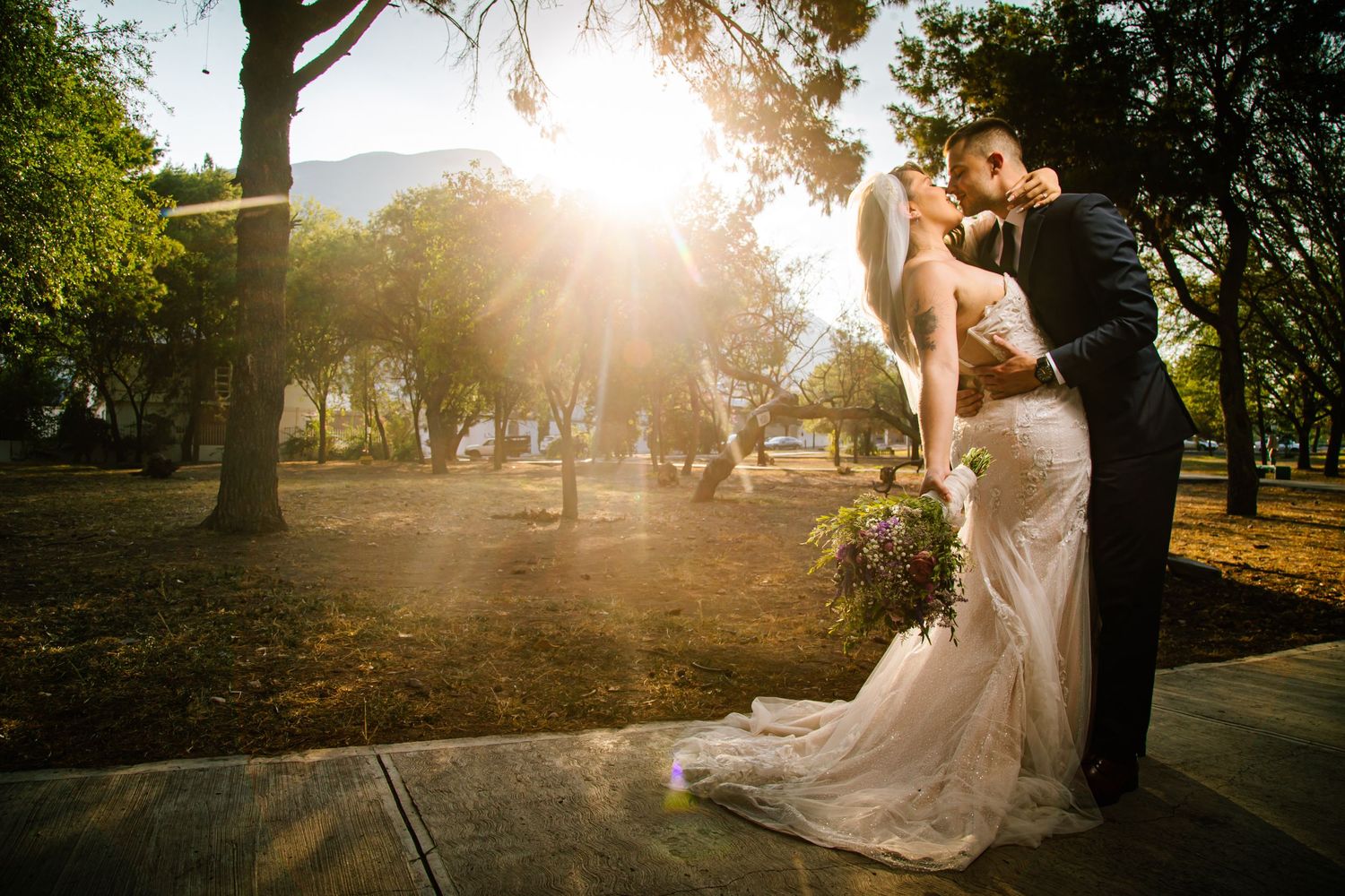 A romantic wedding couple embraces during golden hour in a sunlit park with trees and lens flare effects.
