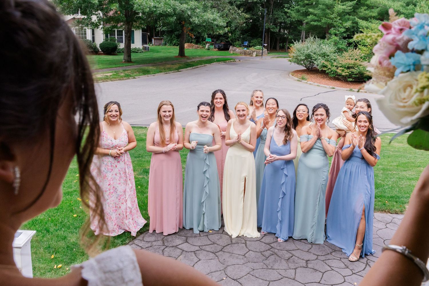 Bridesmaids in pastel colored dresses stand together in a curved line outdoors.