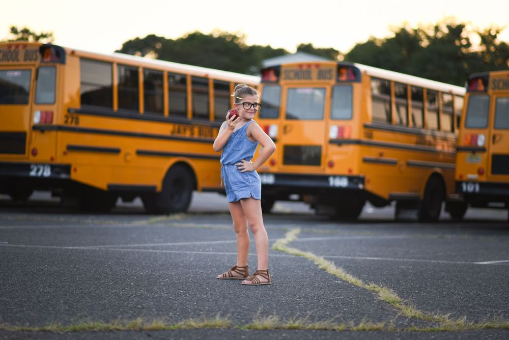 A playful pose of school girl in denim outfit holding a red apple in front of parked yellow school buses. back to school portraitMiddletown