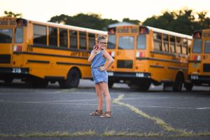 A playful pose of school girl in denim outfit holding a red apple in front of parked yellow school buses. back to school portraitMiddletown