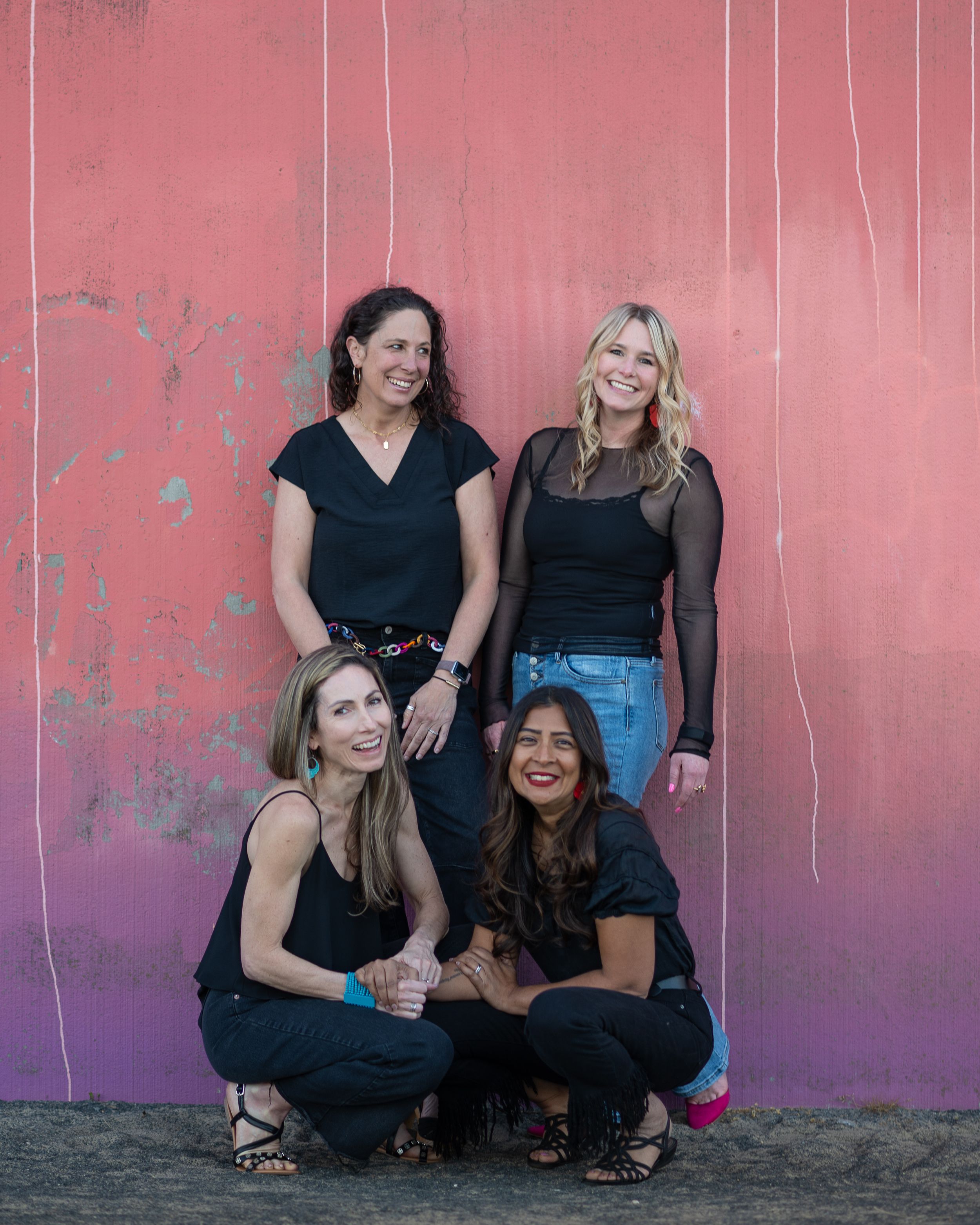Four women in black outfits pose together against a pink wall, two standing and two crouching in front.
