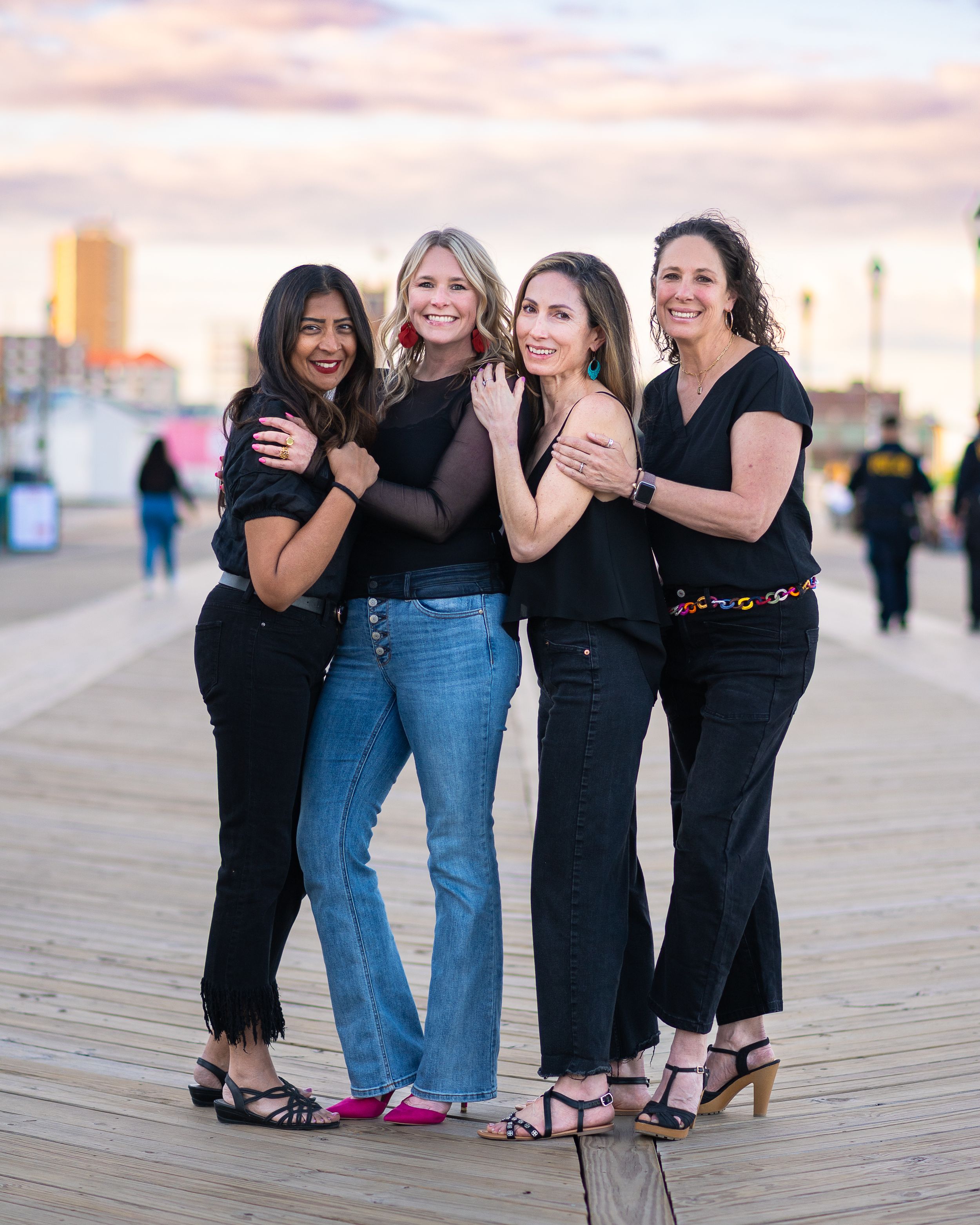 Four women in black outfits pose together smiling on a boardwalk at sunset with a city skyline in the background.