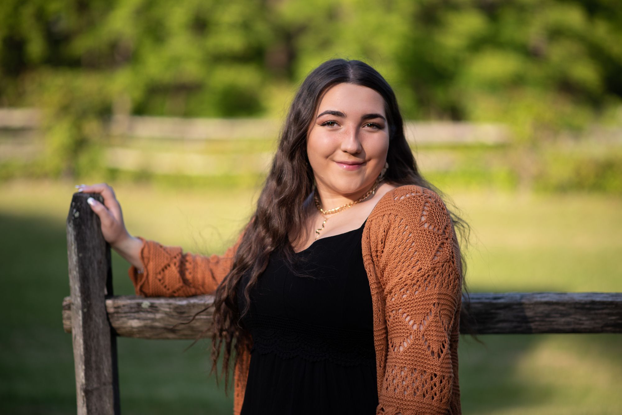 Senior portrait of a girl  in black top and brown cardigan leaning on wooden fence outdoors near Red Bank NJ