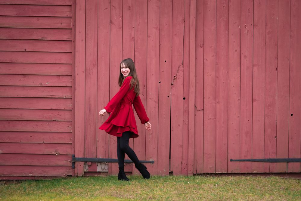 Child portrait of a girl in red dress twirling against a rustic red barn wall in Middletown NJ