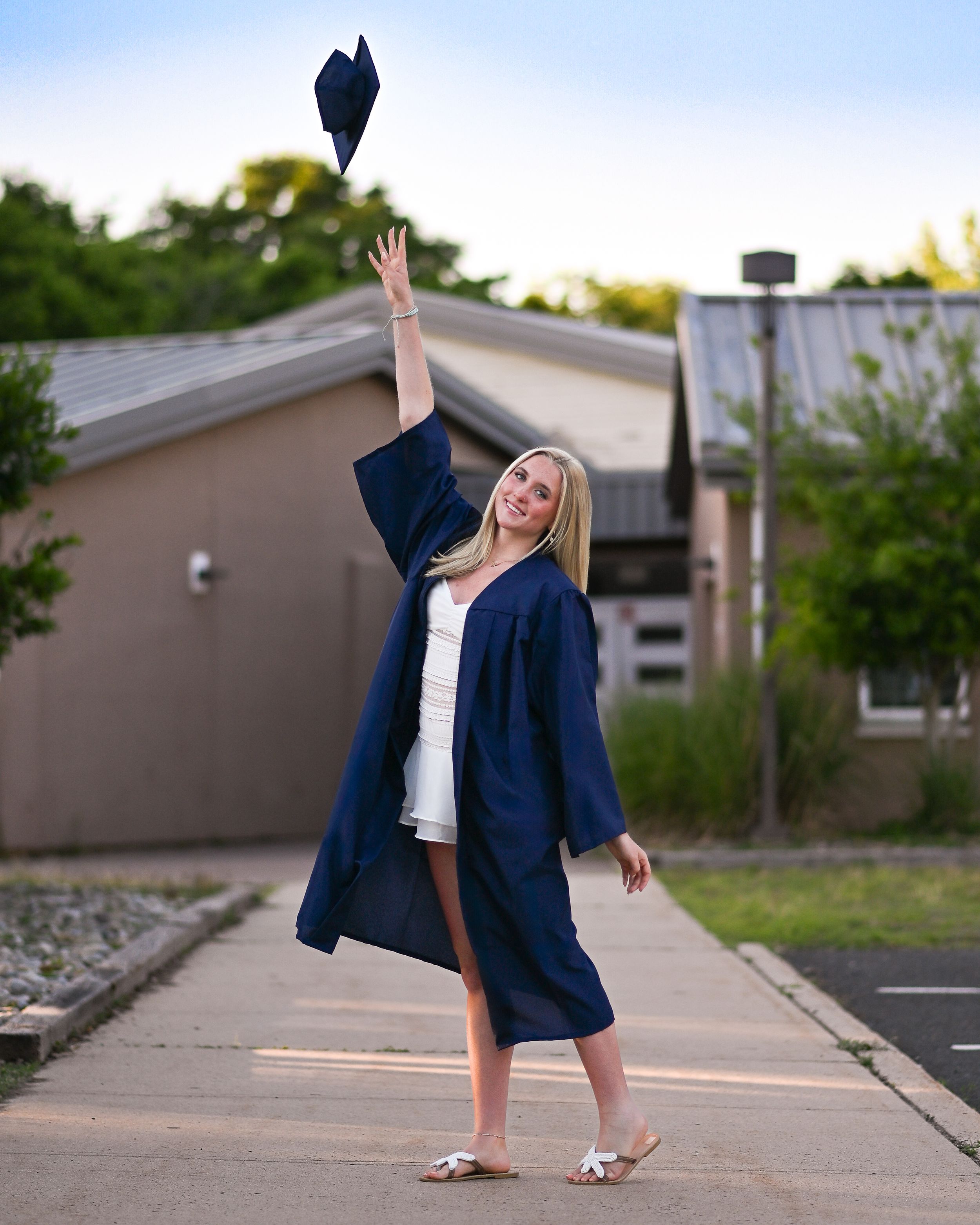 A smiling graduate in navy blue cap and gown tosses her cap in the air outdoors on a sunny evening.