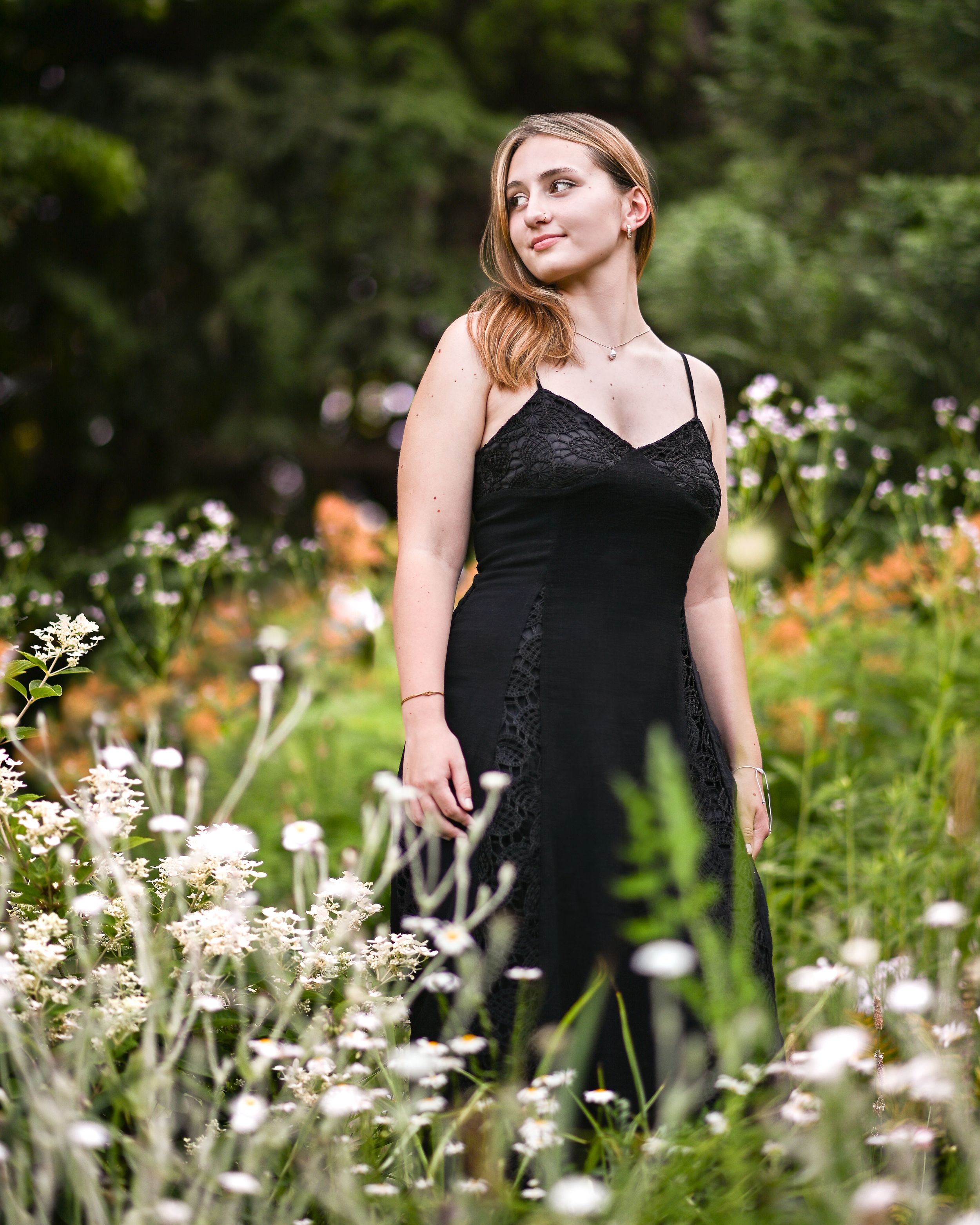 Young woman in black lace dress standing among wildflowers in a lush garden, gazing upward.