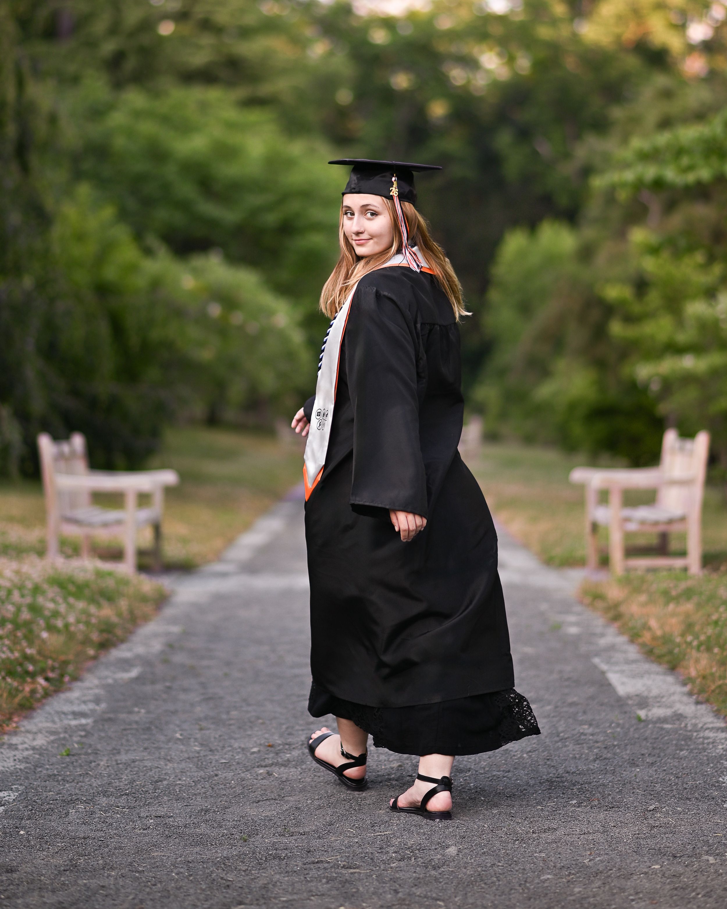 Graduate in black cap and gown poses on a tree-lined path, smiling over her shoulder on graduation day.