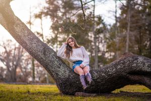 A relaxed senior portrait of a girl sitting on a large curved tree branch in a sunlit park setting in Monmouth County NJ