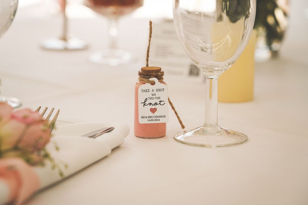 Small pink wedding favor bottle next to wine glasses on white tablecloth.