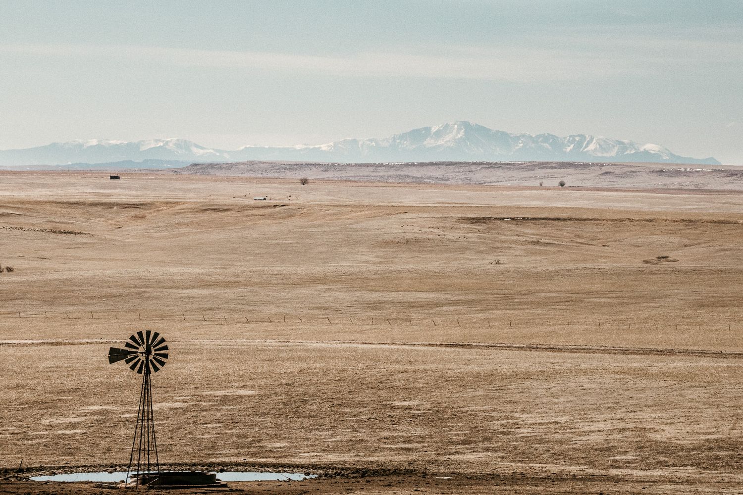 An old weather vane overlooking a large empty field and some farm houses with the Rocky Mountains looming on the horizon.