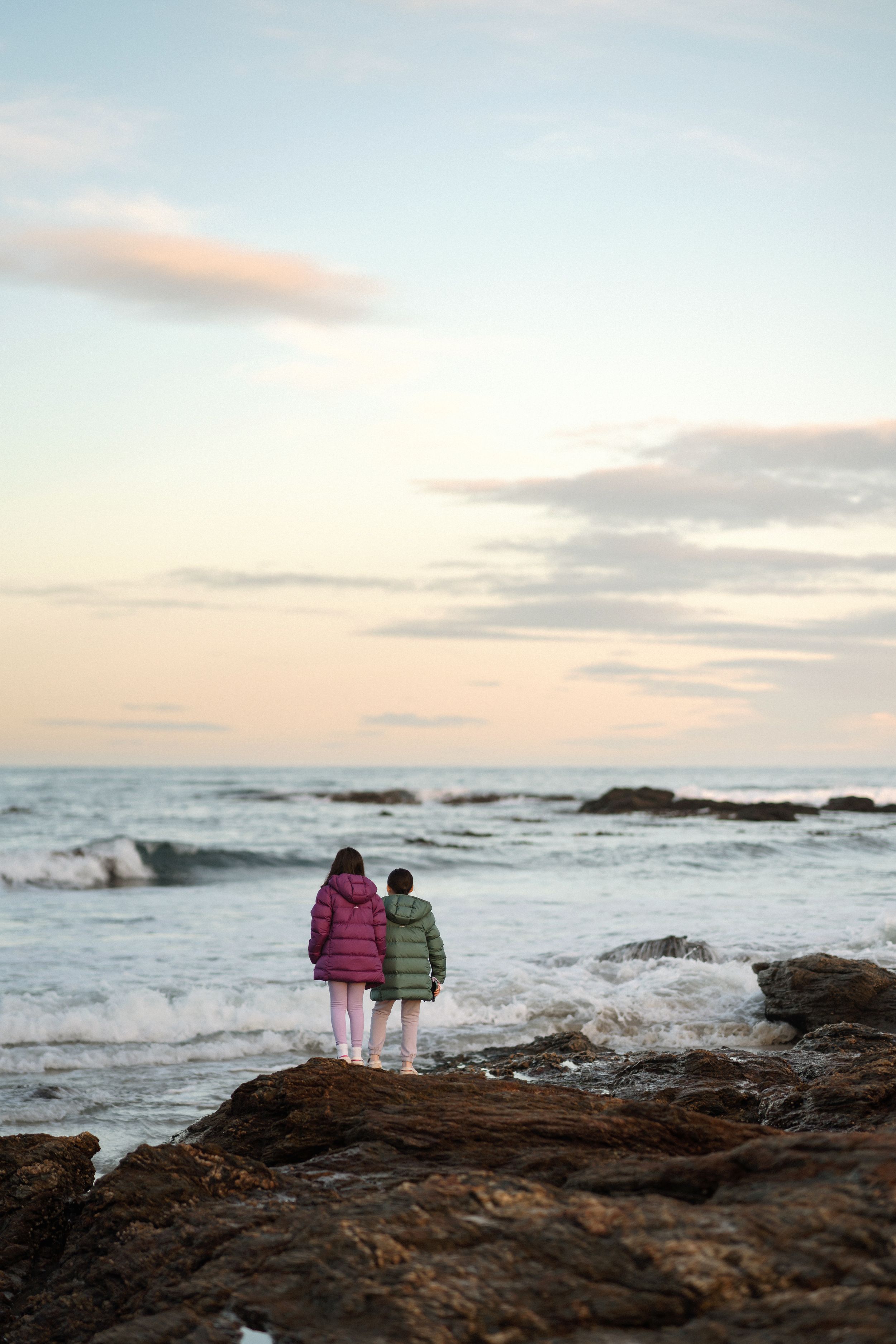 Two children in colorful puffer jackets stand on rocky shore watching ocean waves at sunset.