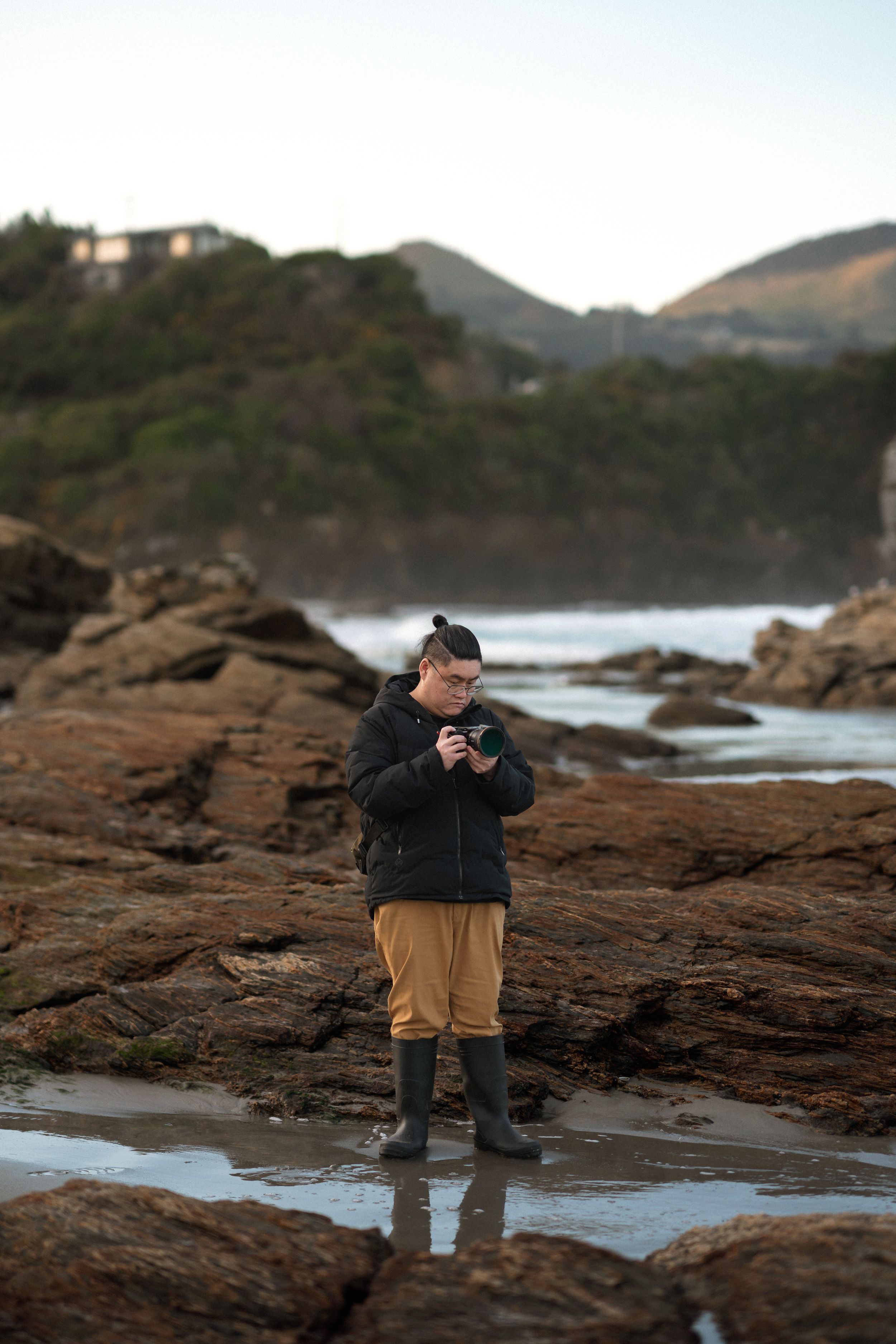 Person standing on rocky coastal shore holding a camera, wearing black jacket and mustard pants, with ocean waves and hills behind.