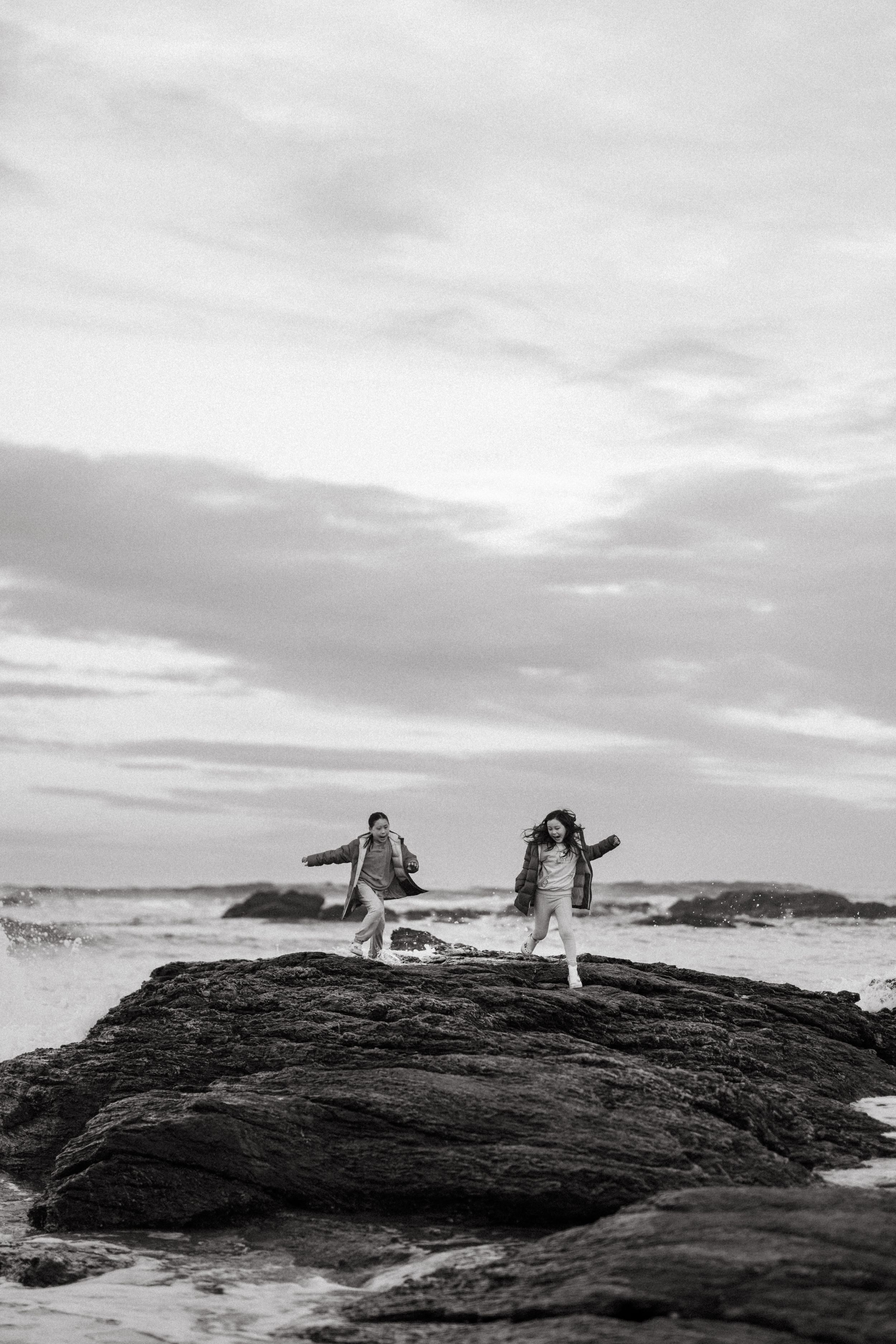 Black and white photo of two people running on rocky coastal cliffs with dramatic ocean waves and cloudy sky.