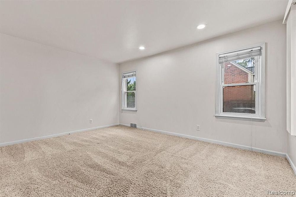 Bright carpeted room with recessed lighting, neutral gray walls, and two windows with natural light.