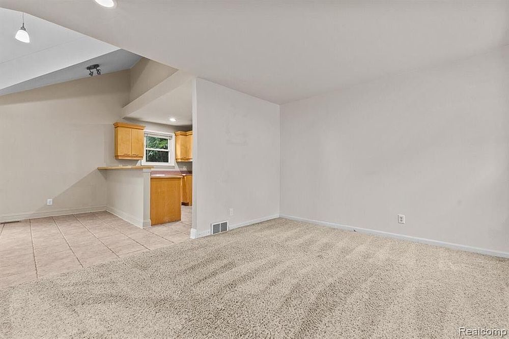 Open basement area with tile and carpet flooring, visible kitchen with wood cabinets in background.