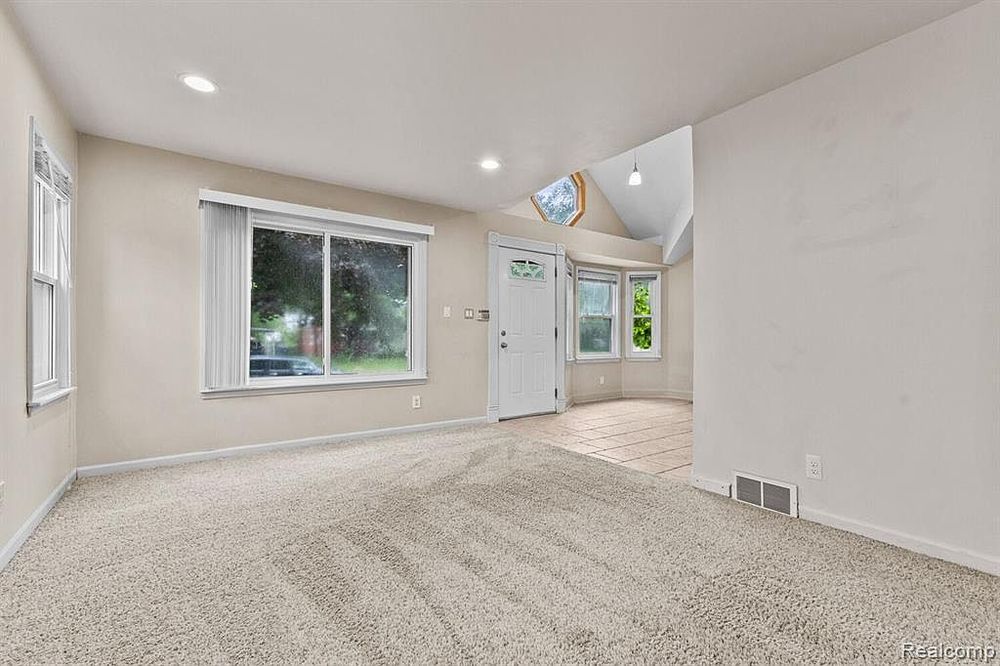 Bright living room with vaulted ceiling, skylight, neutral carpet, and large front windows.