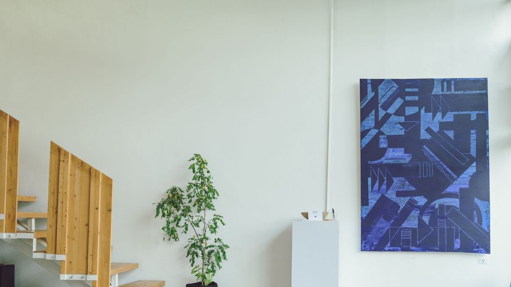 Gallery view showing abstract blue artwork mounted high on white wall near wooden staircase.
