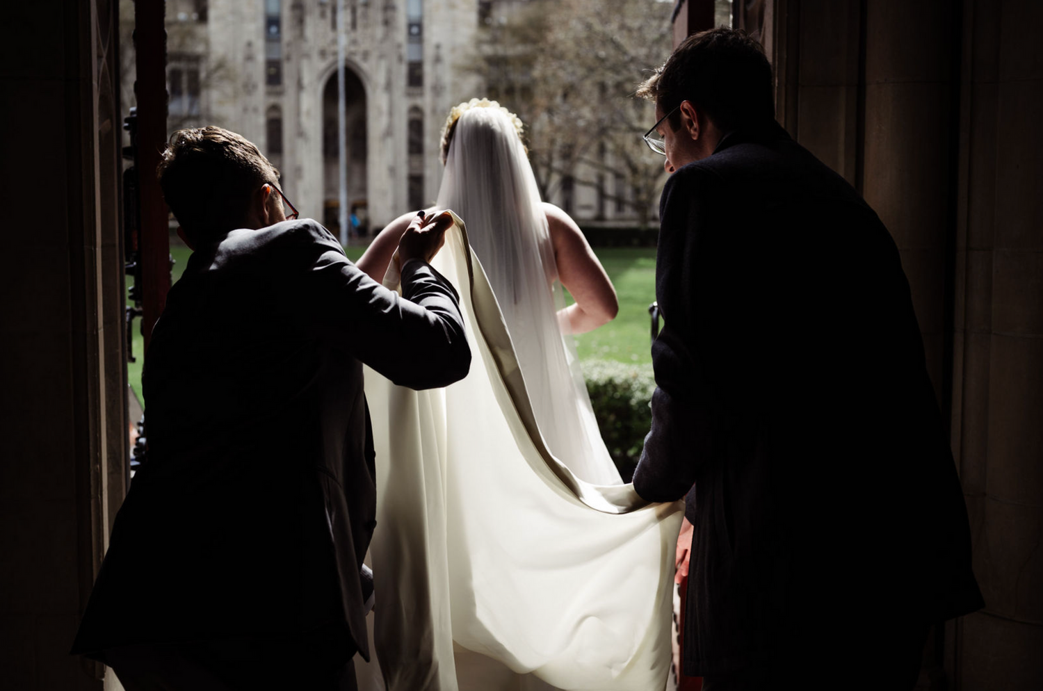 Bride in white veil assisted by two people as she stands in a grand church doorway.