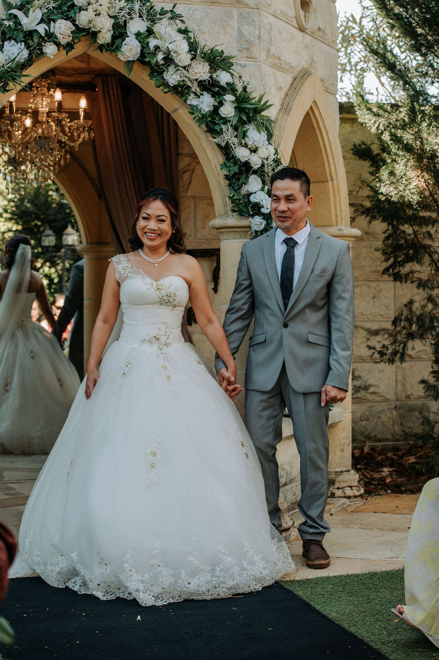 Newlyweds exit under an elegant floral arch decorated with white flowers and crystal chandelier.