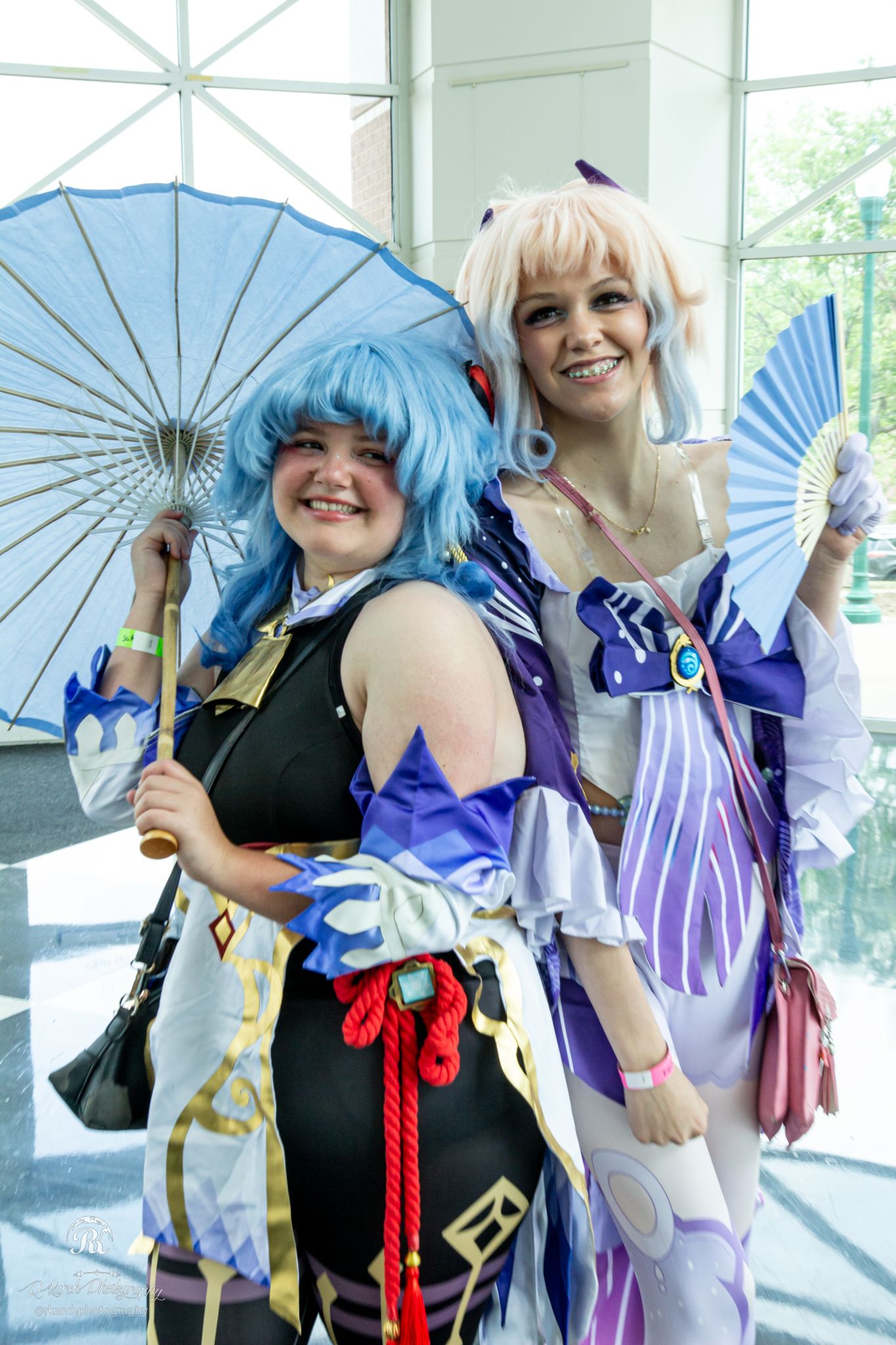 Two cosplayers with decorative fans pose in anime-inspired costumes at a convention.