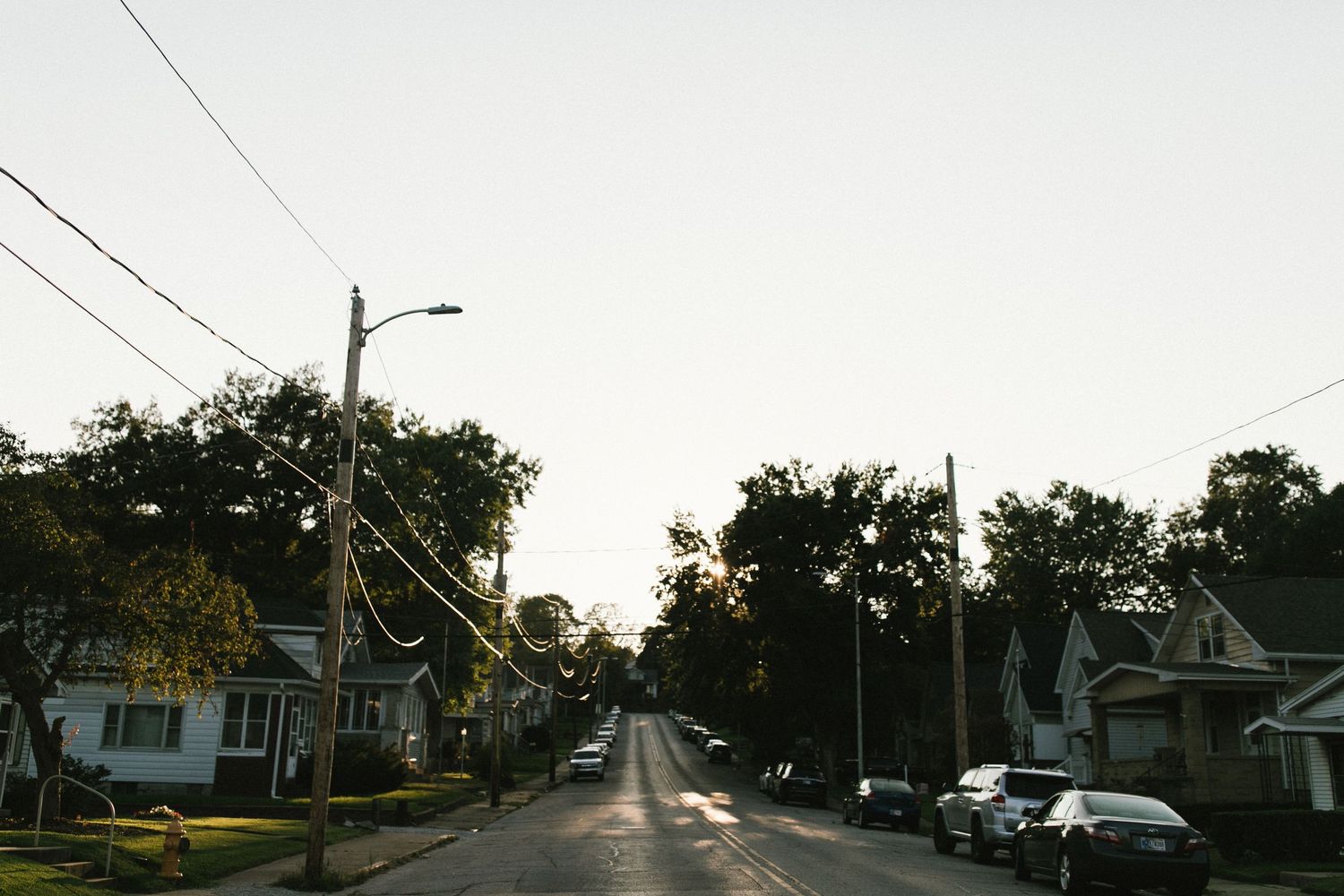 Empty suburban street at dusk with power lines and parked cars.