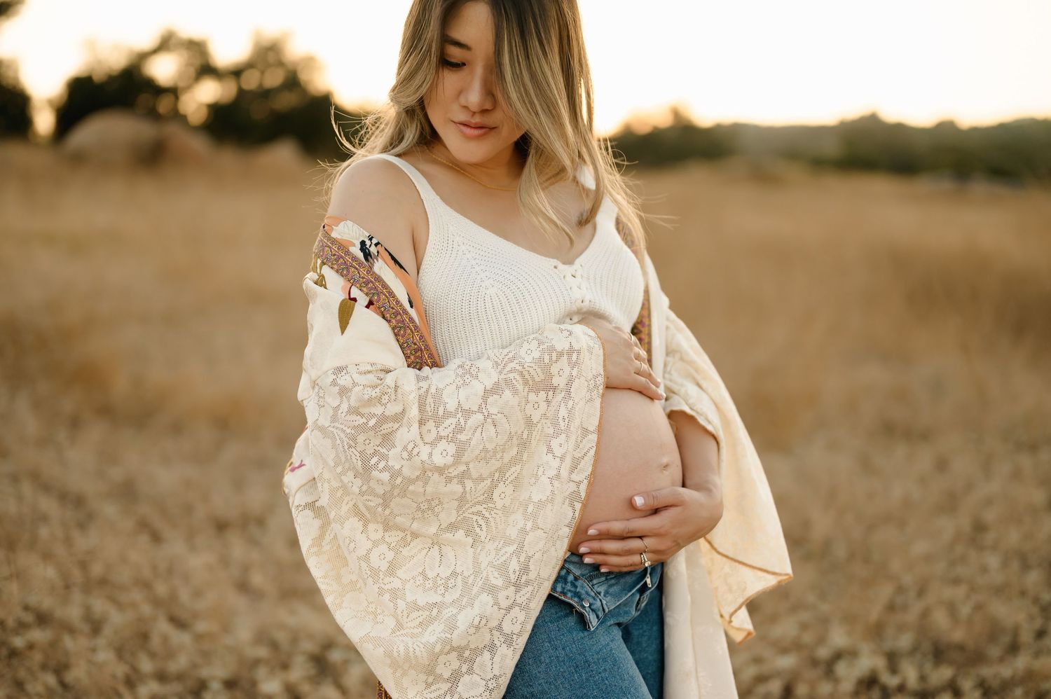 Boho golden grass field maternity photoshoot by Santa Barbara maternity photographer of woman in jeans and white crop top holding bare belly