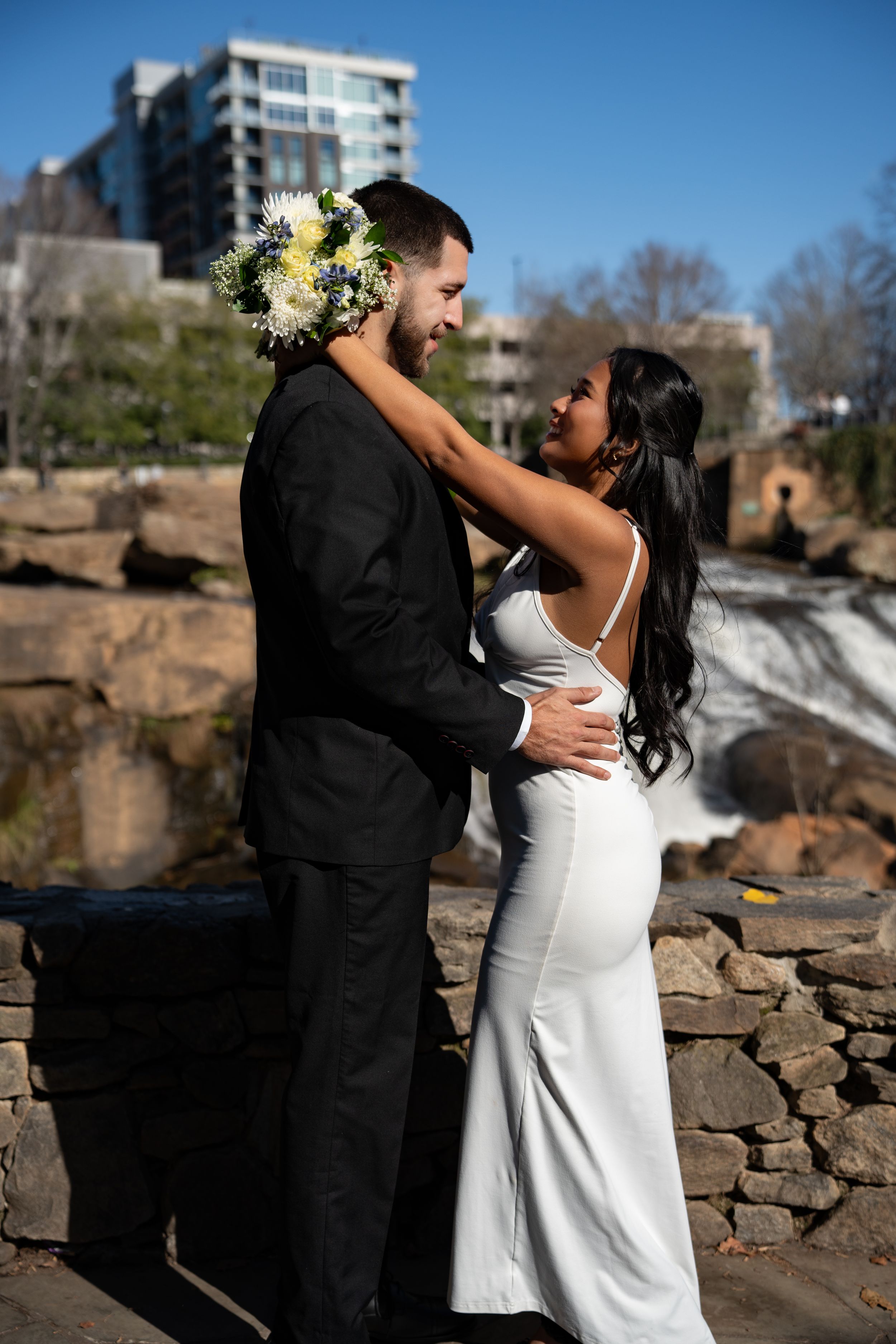 A couple shares a romantic moment on stone steps near a waterfall with the bride wearing a white dress and floral hair piece.