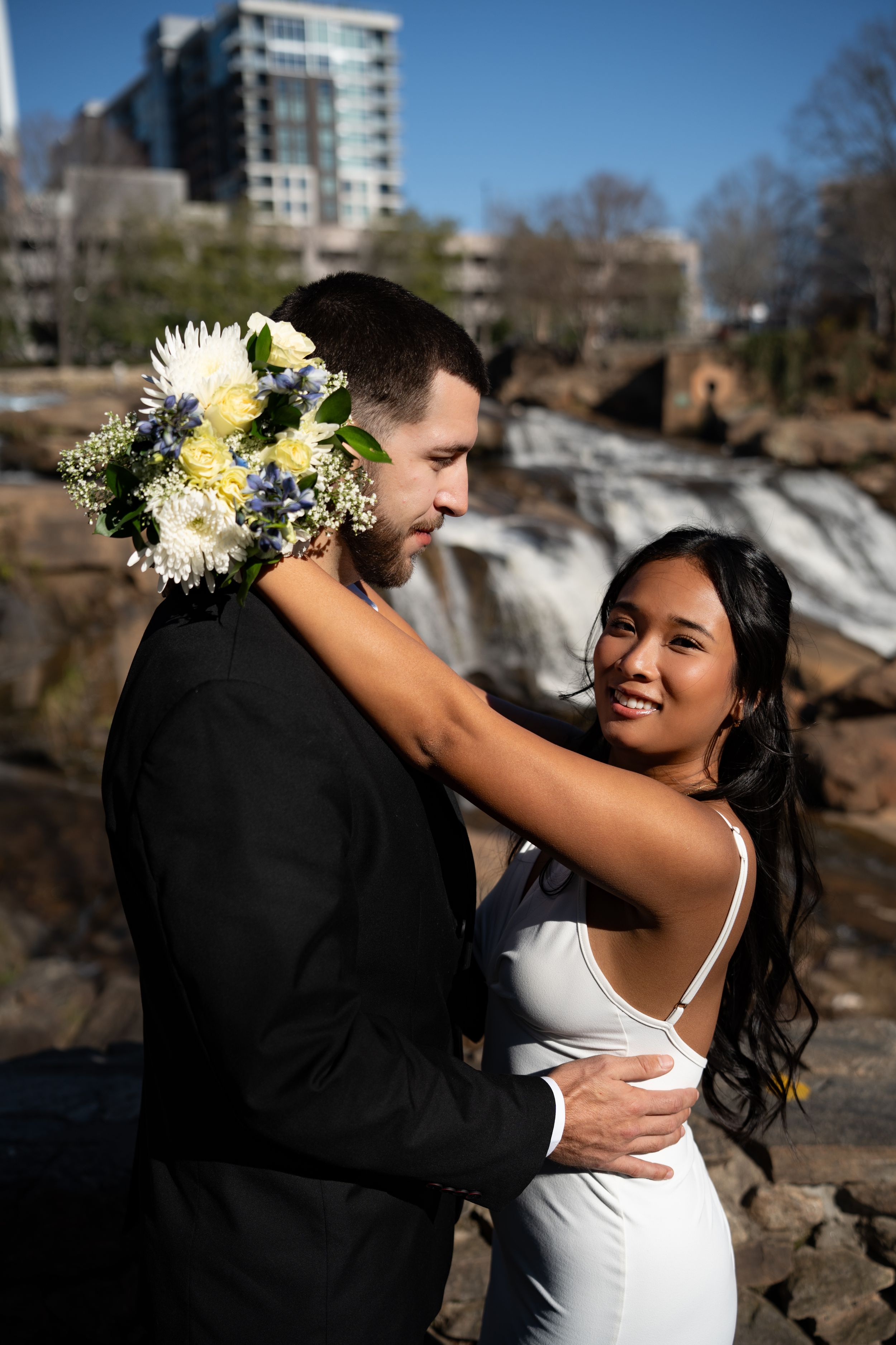 A romantic wedding couple embraces near a scenic waterfall backdrop during their outdoor photo session.